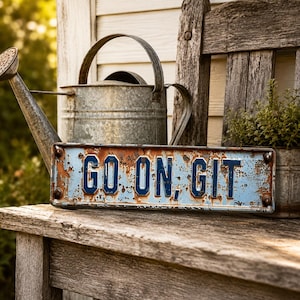 May include: A rectangular metal sign with a distressed light blue finish and dark blue embossed lettering that reads GO ON, GIT. The sign features visible rust along the edges and surface, with four metal rivets securing it at each corner.