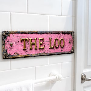 May include: A rectangular wall sign with a distressed pink finish and a dark, weathered border. The sign features raised, metallic gold lettering that spells out the words THE LOO. It is mounted horizontally on a white tiled wall surface.