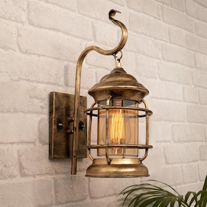 May include: A wall-mounted lantern light fitting with a distressed bronze finish. The lamp features a rectangular backplate, a curved arm with a hook, and a cylindrical glass shade protected by a metal cage. An Edison-style bulb is visible inside.