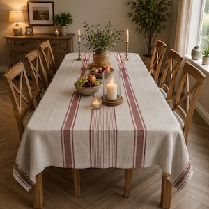 May include: A rectangular dining table covered with a beige linen tablecloth featuring vertical dark red stripes. The table is set with a bowl of fruit, a glass candle holder with a white pillar candle, and two brass candlesticks with lit candles.