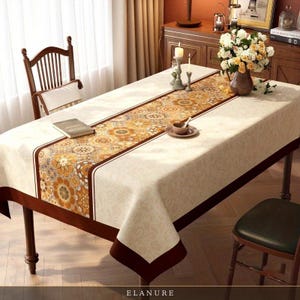May include: A rectangular tablecloth featuring a cream-colored base with a subtle tonal damask pattern and a wide, dark brown border. A central runner displays a repeating circular floral motif in shades of orange, yellow, and brown.