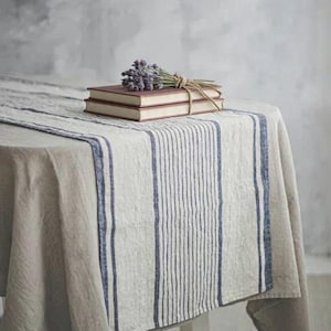 May include: A beige linen tablecloth featuring a central runner with vertical blue stripes. Two stacked rectangular books with red covers are placed on the runner, topped with a small bundle of dried purple lavender tied with twine.