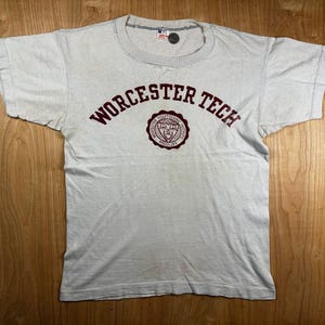 May include: A light gray short-sleeve t-shirt featuring the text WORCESTER TECH in maroon lettering arched over a circular maroon emblem that includes the text WORCESTER POLYTECHNIC INSTITUTE and the Roman numeral MDCCCLXV.