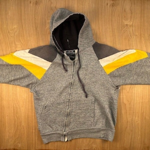 May include: A heather gray zip-up hoodie featuring a black hood lining, long sleeves with diagonal yellow and white stripes across the shoulders, a front zipper closure, two front pockets, and dark gray drawstrings hanging from the hood neckline.
