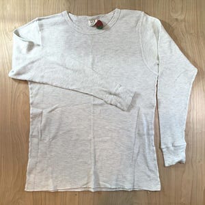 May include: A long-sleeve thermal top in a light grey colour with a waffle-knit texture. The garment features a crew neckline and a small Gap brand label at the inner collar, accompanied by a tiny decorative red strawberry-shaped embellishment.