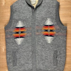 May include: A grey knit waistcoat featuring a shawl collar and a front zip fastening. The garment displays two symmetrical geometric patterns in white, orange, red, and navy blue across the chest. It includes two lower pockets and a Pendleton label.