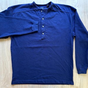 May include: A long-sleeved henley shirt in a solid deep blue colour. The garment features a round neckline with a vertical placket containing four small buttons. The sleeves are finished with ribbed cuffs, and the brand name GAP is visible on the inner neck label.