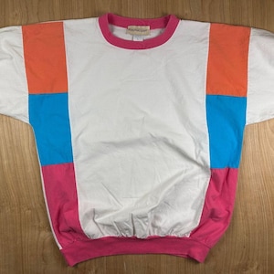 May include: A short-sleeved pullover shirt featuring a white center panel and a round pink neckline. The sides are color-blocked with orange, bright blue, and pink sections. The sleeves and bottom hem are finished with gathered elastic cuffs.