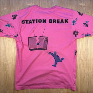 May include: A bright pink short-sleeve t-shirt featuring the text STATION BREAK in black. The shirt displays a graphic of a person throwing an object at a television set, with a repeating pattern of small television and abstract icons on the sleeves.