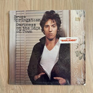 Puede incluir: Una portada de disco de vinilo con Bruce Springsteen vistiendo una chaqueta negra sobre una camiseta blanca de cuello en V. El texto dice Bruce Springsteen Darkness on the Edge of Town e incluye una pegatina blanca INCLUDES BADLANDS.