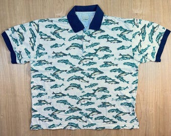 Può includere: Una polo a maniche corte bianca con una stampa all-over di pesci che nuotano in tonalità di verde e blu. Il capo presenta un colletto blu scuro a tinta unita e polsini abbinati, con un'etichetta L.L. Bean sul retro del collo.