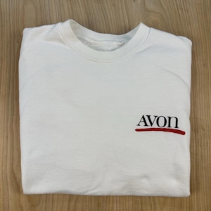 May include: A white crewneck sweatshirt featuring the word Avon printed in black capital letters on the right side of the chest. Below the text is a horizontal red brushstroke line. The garment has long sleeves and a ribbed collar neckline.