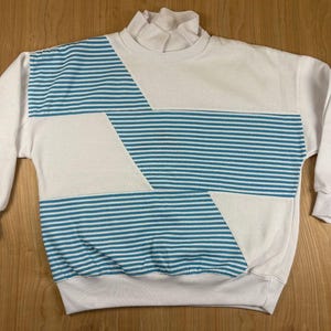 May include: A white long-sleeve sweatshirt featuring a mock neck collar and a geometric pattern of horizontal teal and white stripes. The design consists of alternating solid white sections and striped panels across the front torso.