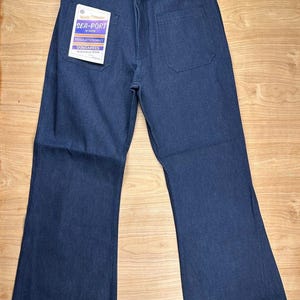 May include: Dark indigo blue denim dungarees featuring a wide-leg cut, a button fly, and two large patch pockets on the front. A white tag attached to the pocket reads World Famous SEA-PORT BY DAVIS REGULATION NAVY DUNGAREES INDIGO BLUE DENIM.