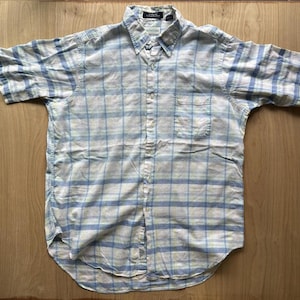 May include: A short-sleeve button-down shirt featuring a light blue, white, and pale green plaid pattern. The shirt includes a standard collar, a single chest pocket on the left side, and a full-length front button closure with white buttons.