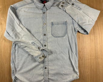 May include: A light-wash blue denim long-sleeve button-down shirt featuring a pointed collar, a single chest pocket on the left side, and metal buttons down the front placket and on the cuffs. The fabric has a faded, worn-in appearance.