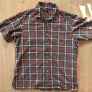 May include: A short-sleeved button-down shirt featuring a plaid pattern in red, green, white, and navy blue. The shirt has a standard collar, a front button placket, and a Polo by Ralph Lauren label on the inside collar area. The fabric is cotton.