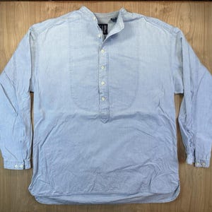 May include: A light blue long-sleeve shirt with a band collar and a partial button-down placket. The shirt features a vertical bib-style panel on the front, long sleeves with buttoned cuffs, and a Gap brand label at the inner neckline.