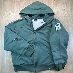 May include: A sage green zip-up hooded sweatshirt with a white thermal lining, featuring a front zipper, two hand pockets, and a patch on the left sleeve displaying a tree graphic and the text Somerset County Park Commission, with visible wear.