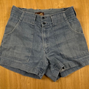 May include: High-waisted denim shorts in a faded blue wash featuring a double-button closure at the waistband, a front zipper fly, and two large patch pockets on the front. The shorts have a structured fit with visible vertical seams.