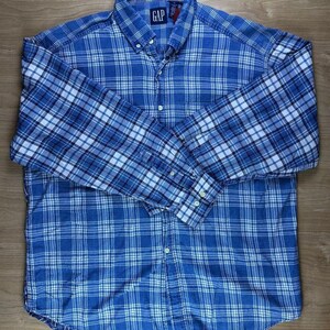 Pode incluir: Uma camisa de manga comprida com botões e um padrão xadrez em azul e branco. A camisa tem um colarinho abotoado, uma carcela frontal com botões e punhos abotoados. Uma etiqueta azul-marinho com o texto branco GAP é visível no interior.
