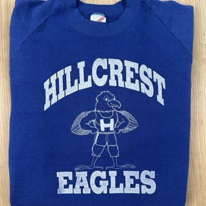 Op de afbeelding: Een koningsblauw sweatshirt met ronde hals en een witte zeefdruk. De tekst HILLCREST staat in een boog boven een adelaar-mascotte die een shirt met de letter H draagt, en het woord EAGLES staat in een rechte lijn onder de mascotte.