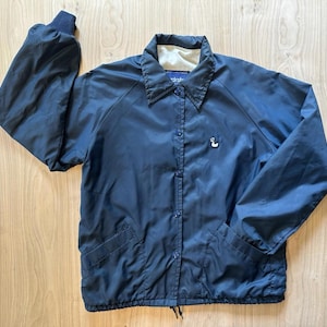 May include: A navy blue windbreaker jacket featuring a pointed collar, long sleeves with elastic cuffs, and a front snap-button closure. The jacket includes two side pockets and a small white embroidered duck logo on the left chest area.