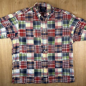 May include: A short-sleeve button-down shirt featuring a multi-colored patchwork plaid pattern. The design consists of various square fabric panels in shades of red, navy blue, white, yellow, and green. It includes a standard collar and buttons.