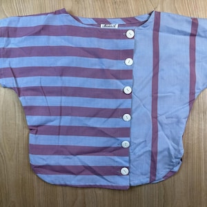 Könnte beinhalten: Eine kurzärmelige Bluse mit geteiltem Design, horizontalen malvenfarbenen und hellblauen Streifen auf der linken Seite sowie vertikalen Streifen auf der rechten Seite. Das Kleidungsstück hat sechs weiße Knöpfe und ein Etikett mit der Aufschrift Mister A.