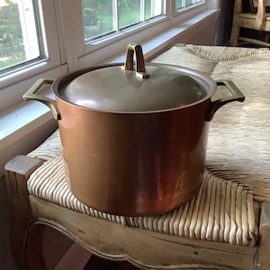 May include: A large, cylindrical copper stockpot with a matching lid and two side handles. The pot features a polished metallic finish with a warm, reddish-brown hue. The lid includes a central handle, and the side handles are securely attached.