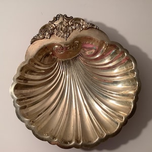 May include: A silver-toned metal dish shaped like a scallop shell. The bowl features deep, radiating fluted grooves and a decorative, ornate embossed scrollwork pattern at the top edge, creating a textured and reflective metallic surface.