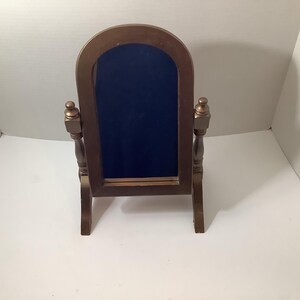 May include: A tabletop mirror with a brown wooden frame and a rounded top. The mirror is supported by two vertical posts with decorative finials on top and stands on two legs. The reflective surface is dark blue, showing a deep reflection.