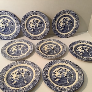 May include: A set of eight round ceramic plates featuring a blue and white Willow pattern. Each plate displays a detailed transfer print of a pagoda, willow trees, a bridge, and birds in flight, surrounded by a decorative geometric border.