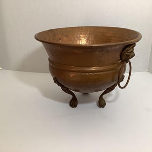May include: A round, hammered copper bowl featuring a wide, flared rim and a smooth, rounded body. The bowl is supported by three ornate, claw-style feet and includes a decorative lion head handle with a metal ring on the side.