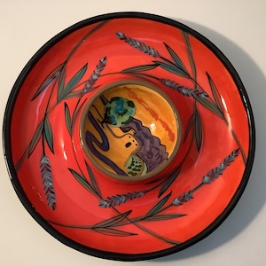 May include: A round ceramic serving dish with a bright orange base and a black rim. The surface features painted lavender sprigs in shades of blue and green. A smaller, circular center bowl displays a painted landscape with a building and tree.
