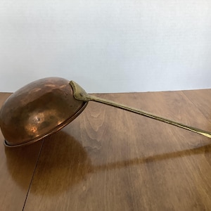 May include: A large, round, hammered copper bowl with a long, slender, polished brass handle attached by a triangular metal bracket. The bowl has a warm, metallic finish, and the handle extends outward, featuring a smooth, tapered design.