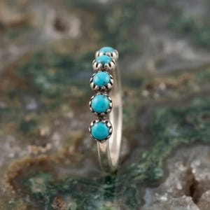 May include: A silver ring featuring five small, round, bright blue turquoise stones set in individual scalloped metal bezels. The stones are arranged in a vertical row along the top of the band, showcasing a polished metallic finish.
