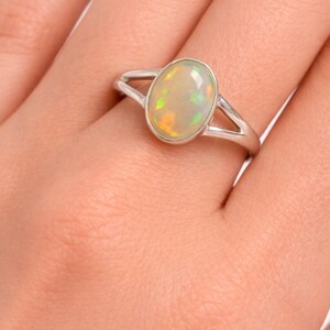 May include: A silver ring featuring a smooth, oval-shaped opal gemstone set in a polished metal bezel. The opal displays iridescent flashes of green, yellow, and orange colour play. The ring band has a split design that tapers towards the setting.
