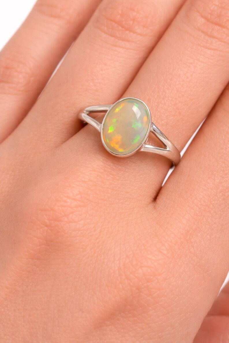 May include: A silver ring featuring a smooth, oval-shaped opal gemstone set in a polished metal bezel. The opal displays iridescent flashes of green, yellow, and orange colour play. The ring band has a split design that tapers towards the setting.