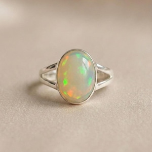 May include: A silver ring featuring a large, oval-shaped cabochon opal gemstone. The opal displays a milky white base with iridescent flashes of green and orange. The ring band is polished silver and splits into two sections near the setting.
