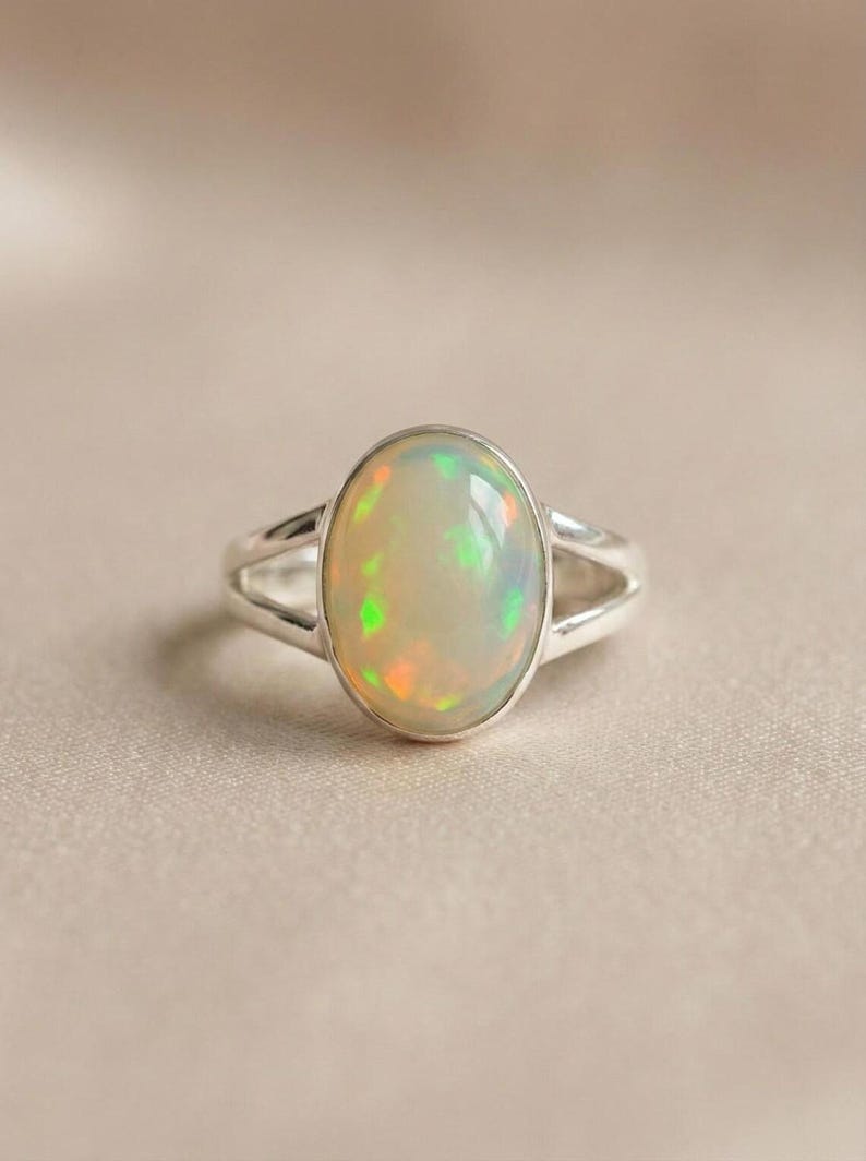 May include: A silver ring featuring a large, oval-shaped cabochon opal gemstone. The opal displays a milky white base with iridescent flashes of green and orange. The ring band is polished silver and splits into two sections near the setting.