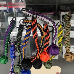 May include: A collection of paracord keychains featuring various braided patterns and colors, including purple, green, blue, orange, black, and tan. Each keychain is finished with a spherical monkey fist knot and a black metal split ring.