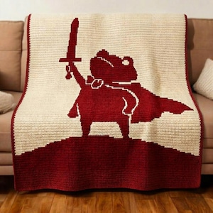 Pode incluir: Uma manta de crochê na cor creme com um design gráfico em vermelho escuro de um sapo vestindo uma capa e segurando uma espada para o alto. A parte inferior da manta é vermelho escuro sólido e toda a peça tem uma borda vermelha escura.