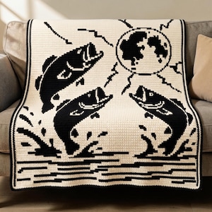 May include: A cream-colored crochet blanket featuring a black graphic design of three jumping fish, water splashes, and a large circular moon with jagged lightning-like rays extending outward, all framed by a solid black border.