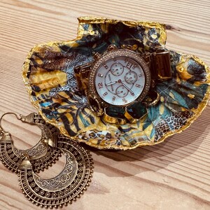 May include: A decorative seashell dish with a gold-painted rim, featuring a patterned interior with leopard print and tropical leaves. Inside the dish sits a gold-tone Michael Kors chronograph watch. Two large, ornate gold-tone hoop earrings lie nearby.