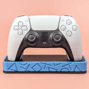 Op de afbeelding: Een wit en zwart draadloze videogamecontroller rustend op een rechthoekige lichtblauwe standaard. De standaard is voorzien van reliëf geometrische vormen, waaronder driehoeken, cirkels, vierkanten en kruisen op de voorzijde.