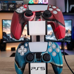 May include: A white plastic dual controller stand featuring the PS5 logo on the base. The stand holds two wireless game controllers, one with a red and black marbled pattern and the other with a blue and black marbled pattern, both featuring white buttons.