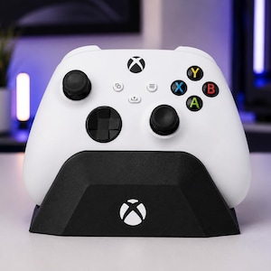 Op de afbeelding: Een witte draadloze videogamecontroller met zwarte analoge sticks en een zwarte richtingspad, voorzien van het Xbox-logo en kleurrijke actieknoppen. De controller rust in een zwart oplaadstation dat ook het witte Xbox-logo weergeeft.