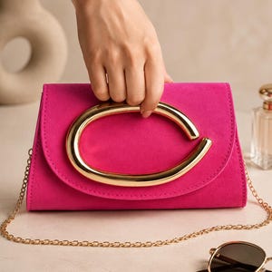 Könnte beinhalten: Eine leuchtend fuchsiafarbene Clutch mit glatter, matter Textur und einem großen, polierten, goldfarbenen ovalen Griff, der mittig auf der Frontklappe sitzt. Die Tasche verfügt über einen feinen, goldfarbenen Metallketten-Schulterriemen.