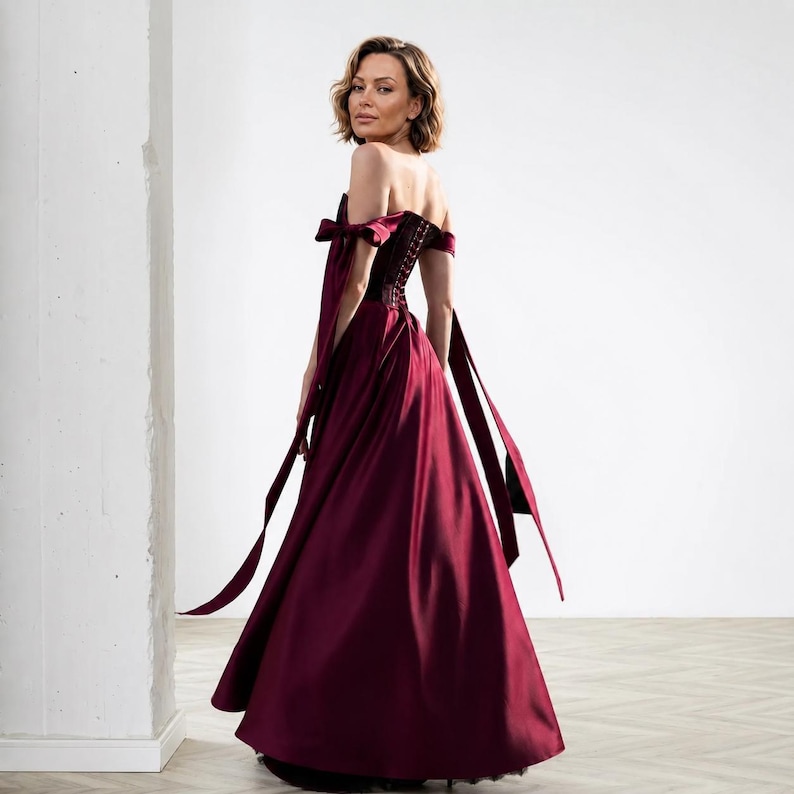 May include: A floor-length burgundy satin gown featuring an off-the-shoulder neckline with tied ribbon sleeve details, a structured corset-style bodice with visible back lacing, and a full, flowing skirt that extends to the floor.